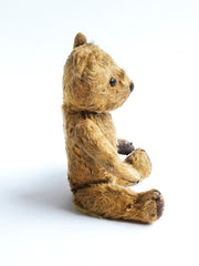 A charming and much sort after vintage 1920s teddy bear. Made from a classic shade of gold mohair, he has been much loved. His neat paw pads are made of brown leather and each has three embroidered claws. He has amber coloured eyes; his nose and mouth are embroidered; his head and arms rotate, and he has swivel jointing to his stout legs. He is stuffed with his original wood-wool fill.