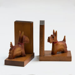 A charming pair of 1930s carved wood scottie dog bookends. Each dog has a painted red collar and tongue, and is mounted on an oak platform with an oak support modelled as a book. Scottie dogs were all the rage in the 1930s: as pets with box-clipped muzzles; as stylised Art Deco Bakelite brooches and clasps; adorning greetings and postcards; as ornaments, modelled in china, metal and wood; and as bookends - such as this dear little pair.