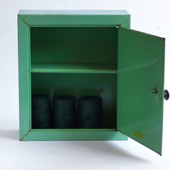 A 1930s steel wall-mounted bathroom cabinet with its original apple green paint. It has a hinged door with arched mirror insert and an internal shelf, and the door is operated by pulling on a Bakelite knob that clips shut when closed.