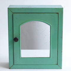 A 1930s steel wall-mounted bathroom cabinet with its original apple green paint. It has a hinged door with arched mirror insert and an internal shelf, and the door is operated by pulling on a Bakelite knob that clips shut when closed.