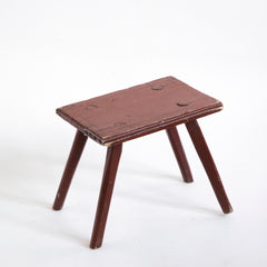 A Romanian folk art small side table or stool with stick legs and with its original brick-red painted finish, and a charming small sheet-metal repair to one corner of its top.