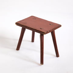 A Romanian folk art small side table or stool with stick legs and with its original brick-red painted finish, and a charming small sheet-metal repair to one corner of its top.