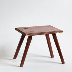 A Romanian folk art small side table or stool with stick legs and with its original brick-red painted finish, and a charming small sheet-metal repair to one corner of its top.