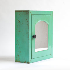 A 1930s steel wall-mounted bathroom cabinet with its original apple green paint. It has a hinged door with arched mirror insert and an internal shelf, and the door is operated by pulling on a Bakelite knob that clips shut when closed.