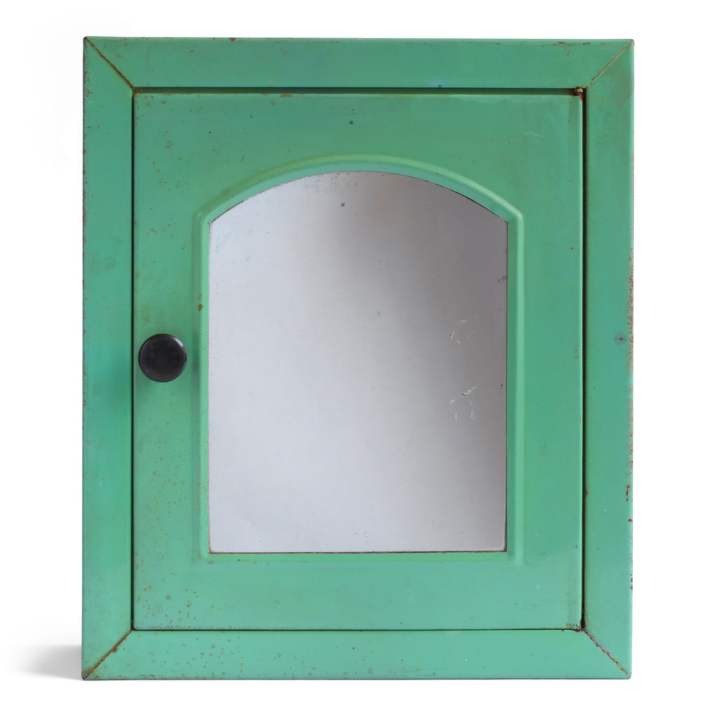 A 1930s steel wall-mounted bathroom cabinet with its original apple green paint. It has a hinged door with arched mirror insert and an internal shelf, and the door is operated by pulling on a Bakelite knob that clips shut when closed.