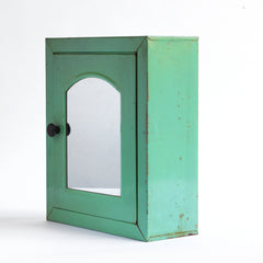 A 1930s steel wall-mounted bathroom cabinet with its original apple green paint. It has a hinged door with arched mirror insert and an internal shelf, and the door is operated by pulling on a Bakelite knob that clips shut when closed.