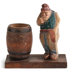 A mid twentieth century naive carved wooden figure of a contemplative rural man standing next to an open barrel.