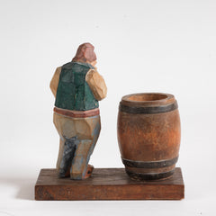 A mid twentieth century naive carved wooden figure of a contemplative rural man standing next to an open barrel.