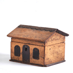 A vintage naive model wooden house savings box that resembles a Scottish bothy, with metal trim to its roof, along with metal windows and front door.