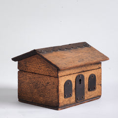 A vintage naive model wooden house savings box that resembles a Scottish bothy, with metal trim to its roof, along with metal windows and front door.