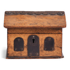 A vintage naive model wooden house savings box that resembles a Scottish bothy, with metal trim to its roof, along with metal windows and front door.