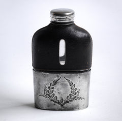 A quality Edwardian silver plated, glass and black leather hip flask with a screw-cap top, and beautiful leaf decoration etched into the silver plate. The clear glass body is half covered with leather, the lower half has a detachable silver plated cover that also acts as a cup.