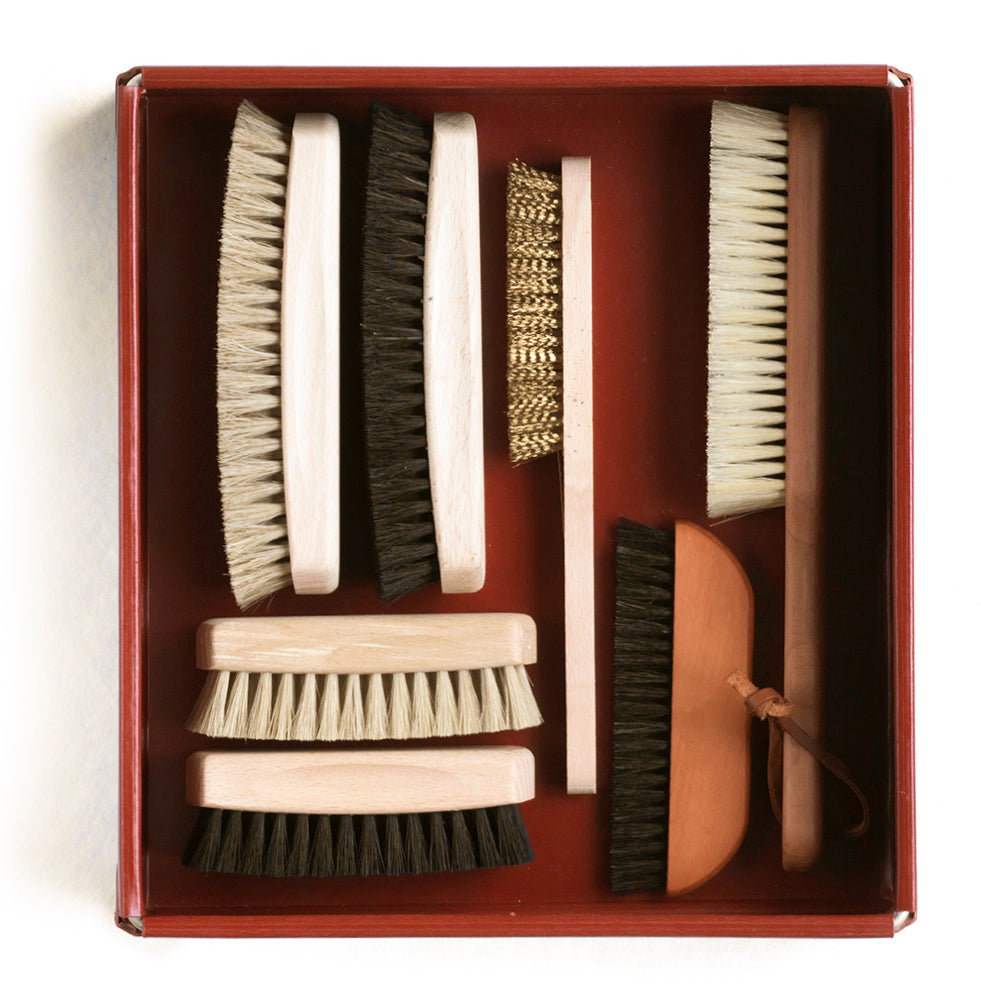 A collection of various brushes and cleaning tools displayed neatly in a ox blood red box. The items include a clothes brush, a shoe brush, and a brass wire brush among others.