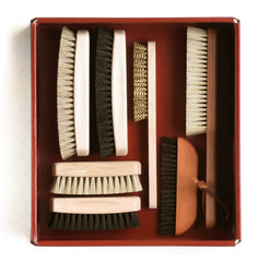A collection of various brushes and cleaning tools displayed neatly in a ox blood red box. The items include a clothes brush, a shoe brush, and a brass wire brush among others.
